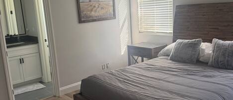 A bedroom with a bed, a nightstand, a window with blinds, a mirror, and a door leading to a bathroom.