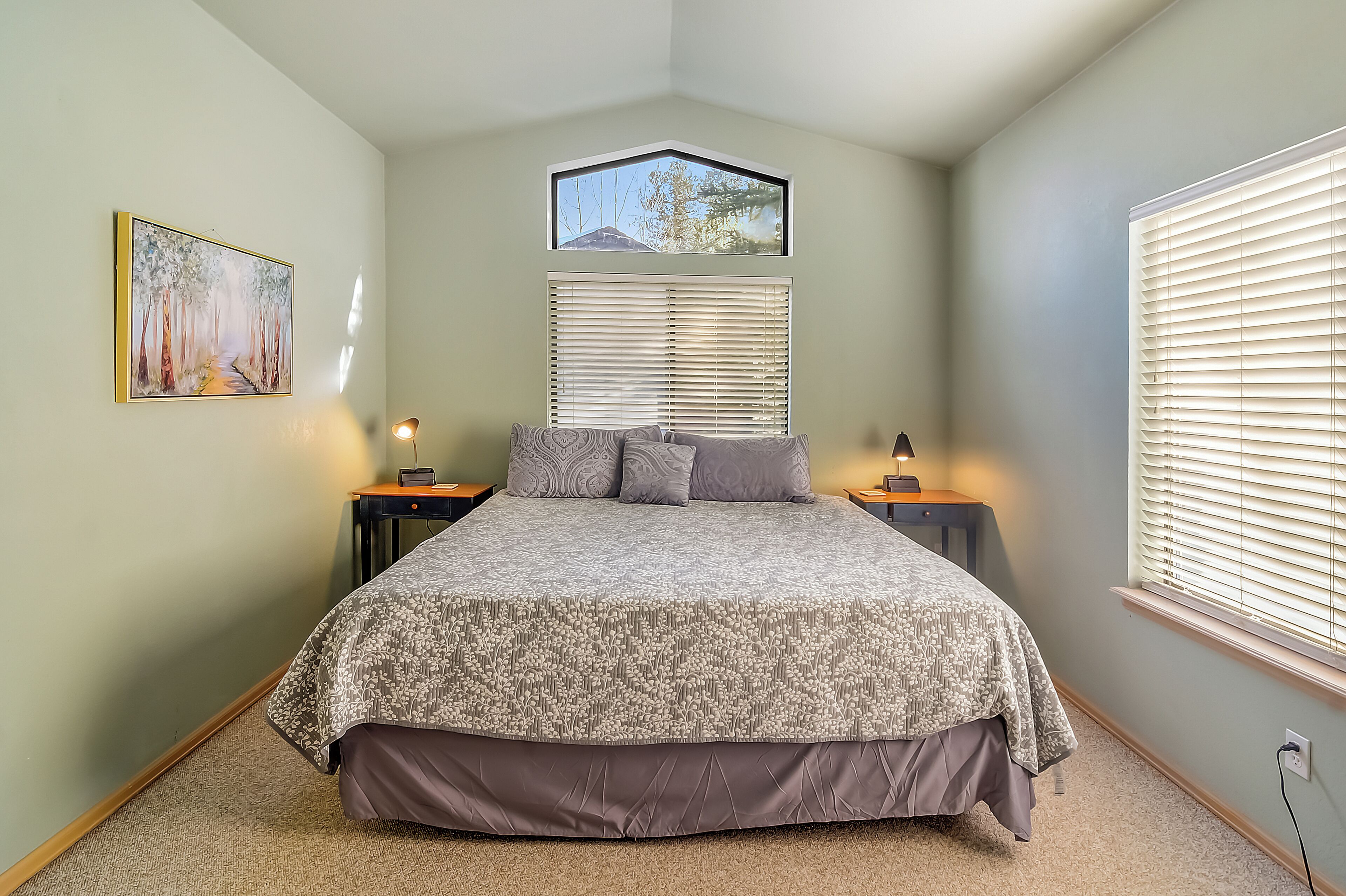 A bedroom with a bed, two bedside tables, a window with blinds, a wall-mounted picture, and a small skylight.