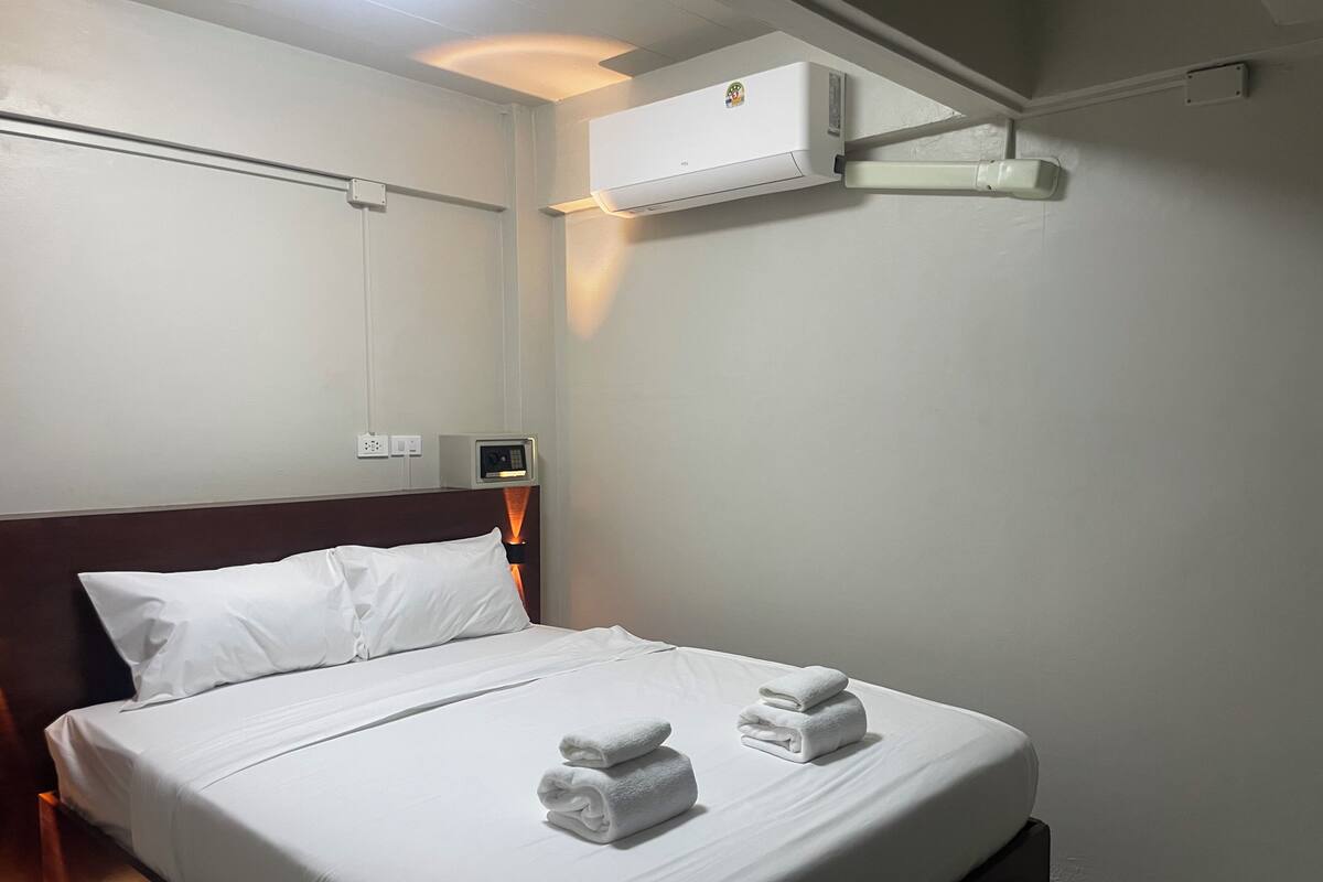 Habitación de hotel con cama, ropa de cama blanca, dos toallas, unidad de aire acondicionado, panel de control y toma de corriente.