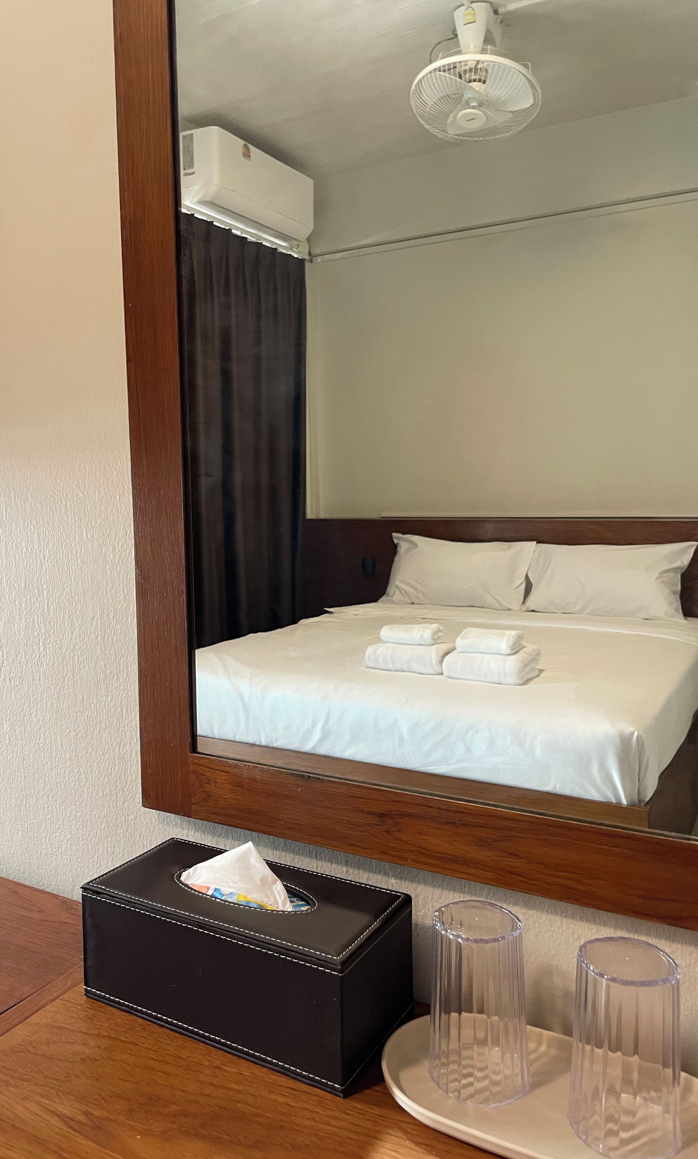 A hotel room with a bed, a bedside table with a tissue box and glasses, and a mirror reflecting the room.
