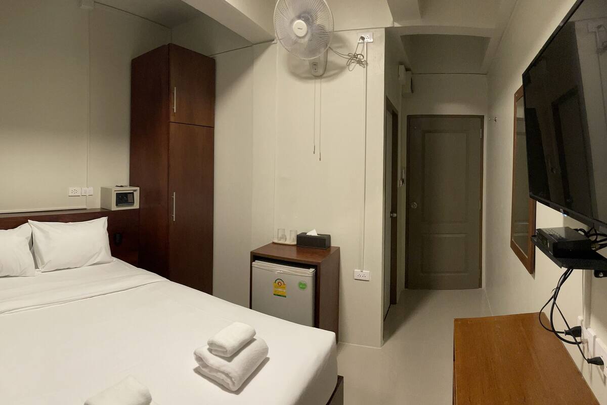 Una habitación de hotel con cama, televisión, ventilador, una mesita y armario.