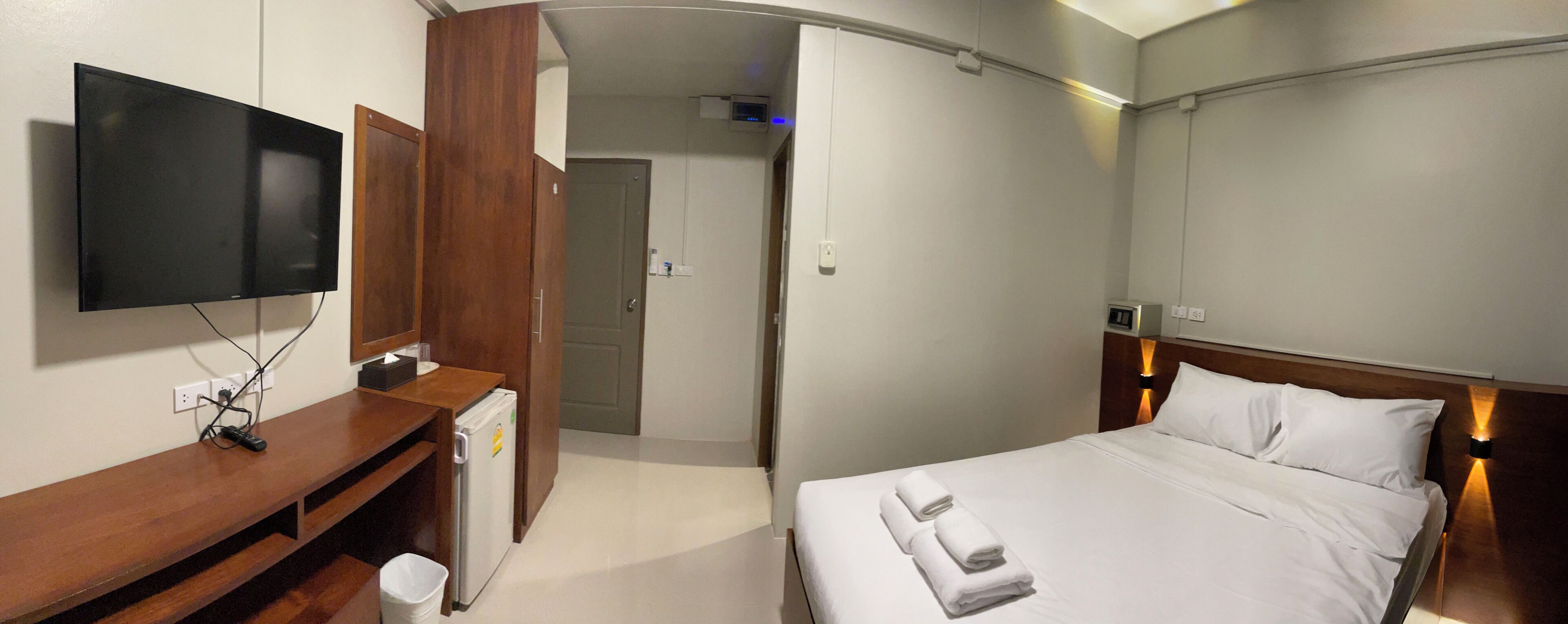 A hotel room with a bed, a TV mounted on the wall, a wooden desk, and a refrigerator.