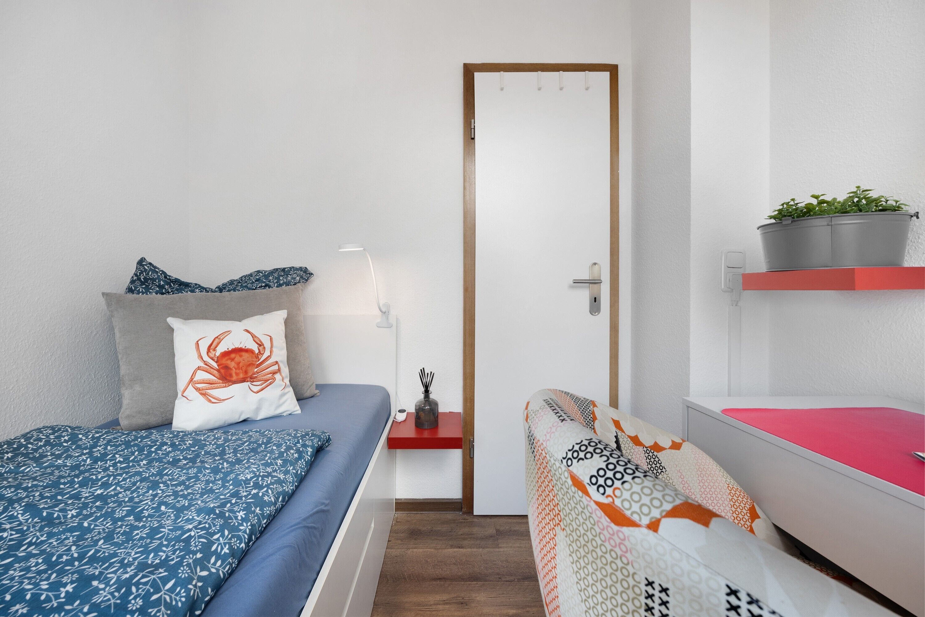 Une chambre à coucher avec un lit, une table de chevet, une étagère rouge ornée d’une plante et une porte.