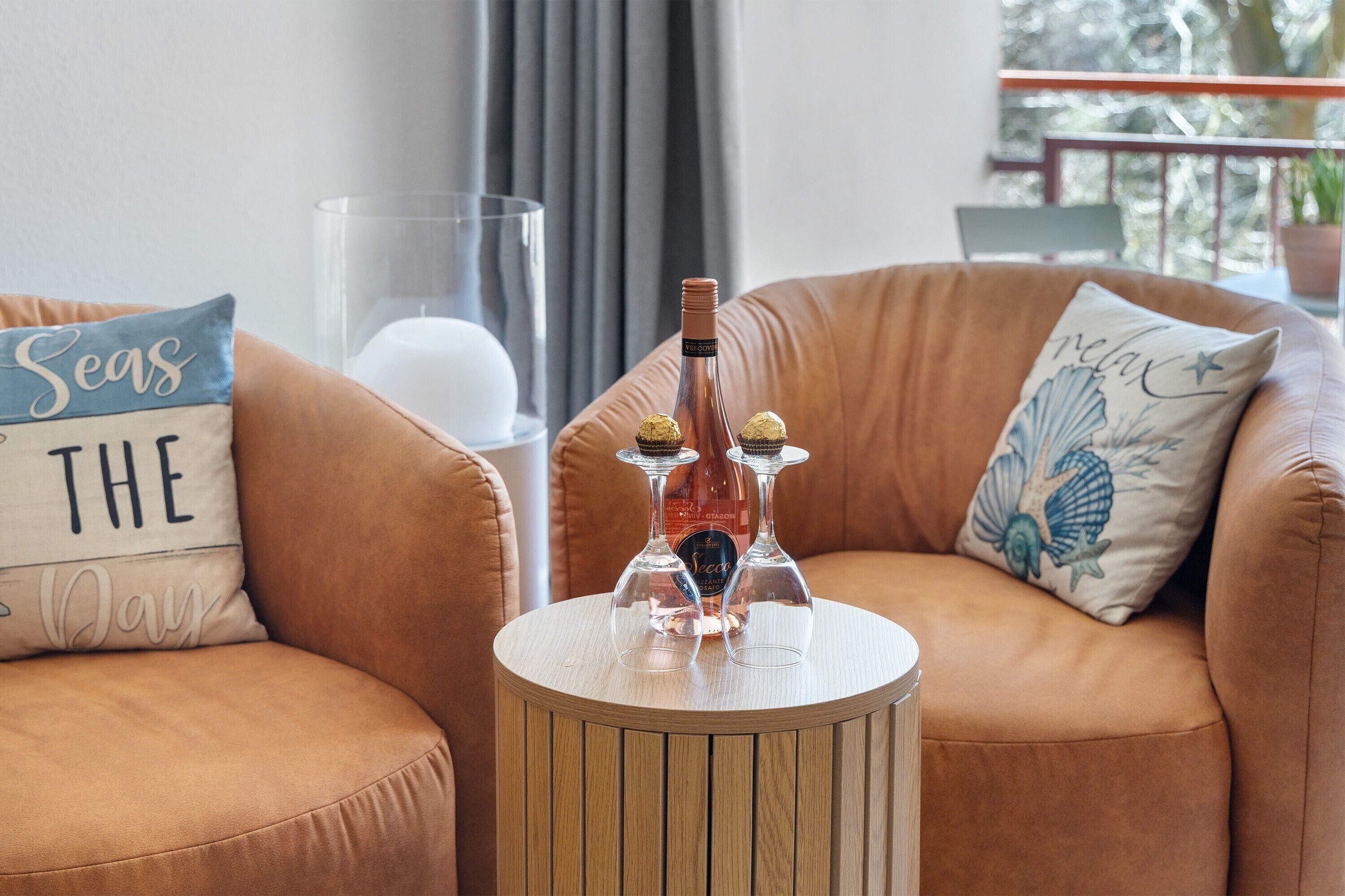 A brown leather sofa with a round wooden side table, a glass candle holder, two wine glasses, and a bottle of wine.