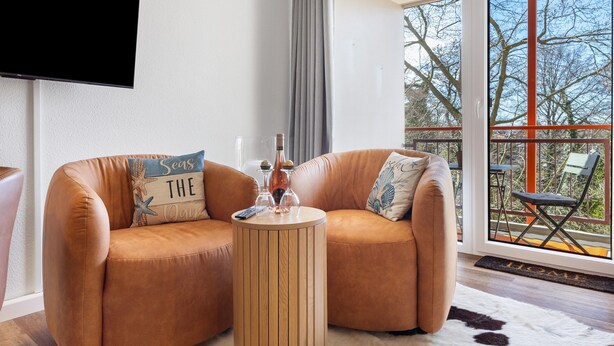 A modern living room with a brown leather sofa, a wooden coffee table, and a large window with a view of trees. - Apartment in the 'Krabbenkiste' Hotel with Sea View, Shared Garden, and Wi-Fi (Wunstorf)