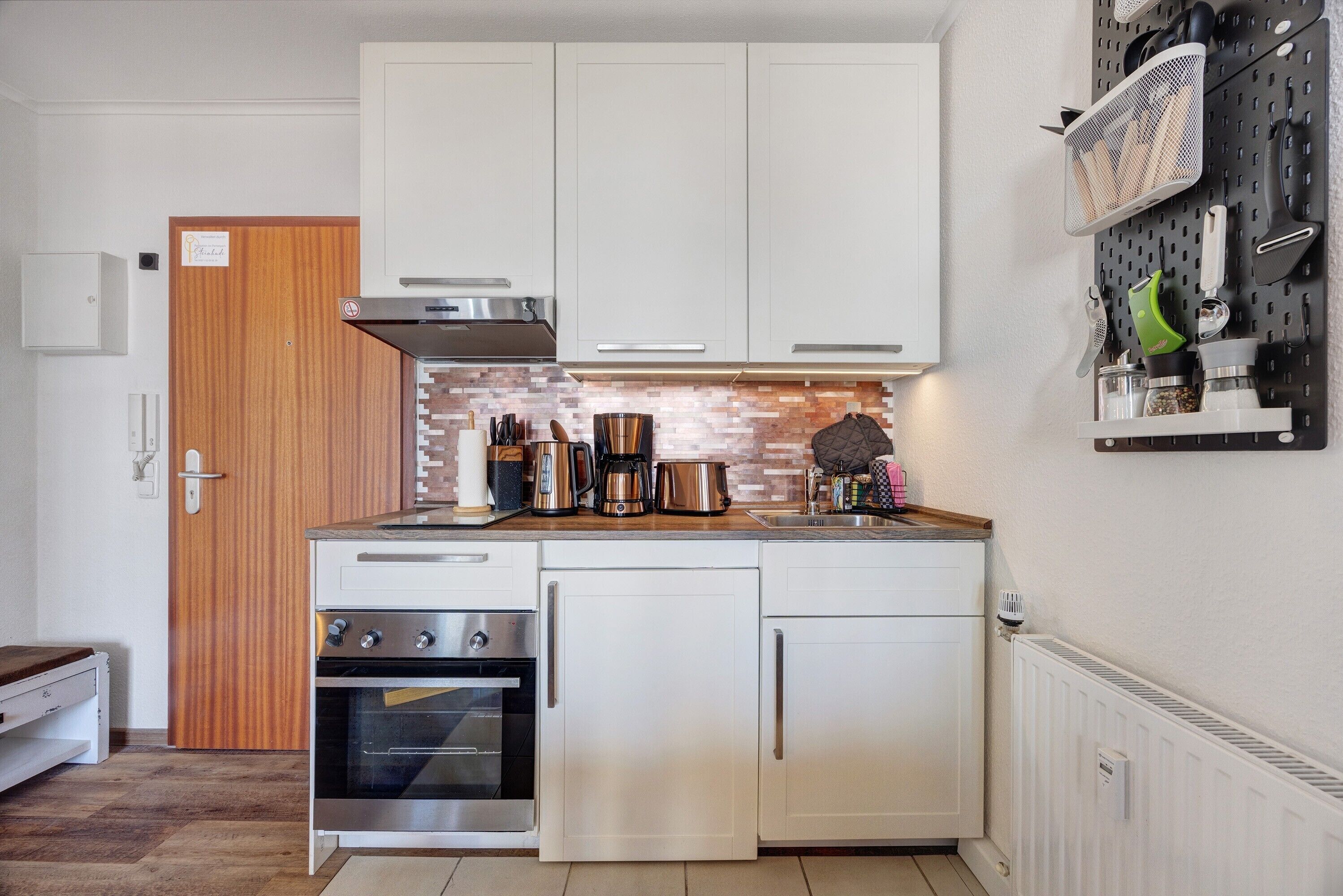 A compact kitchen with white cabinets, a stainless steel oven, a coffee maker, and a pegboard with kitchen utensils.