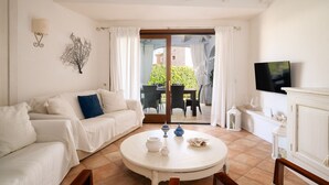 Een kamer met een witte bank, een ronde tafel met een theepot en kopjes, een televisie aan de muur en uitzicht op een tuin door een glazen deur.