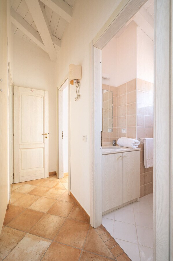 Un pasillo que conduce a un baño con suelo de baldosas, una puerta blanca y un espejo sobre el lavabo.