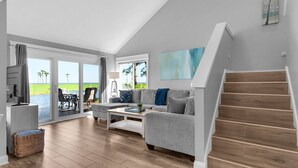 A spacious living room with a large sliding glass door leading to a balcony, a grey sofa, a coffee table, and a staircase. - 🌞Bayfront | Pet Friendly | Golf Cart | Pool | Fairways 274- Sea Ya Later🌞 (Miramar Beach)