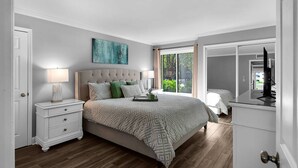 A bedroom with a large bed, a nightstand, a dresser, a window with curtains, and a painting on the wall. - 🌞Bayfront | Pet Friendly | Golf Cart | Pool | Fairways 274- Sea Ya Later🌞 (Miramar Beach)