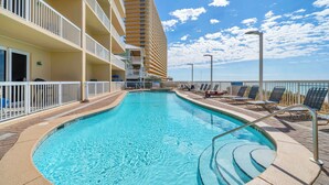 Ein Pool mit Blick auf ein hohes Gebäude, ein Balkon mit Liegestühlen und ein klarer Himmel.