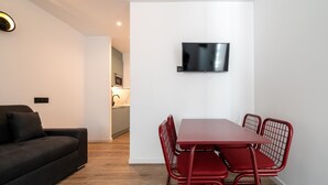 A modern living room with a dark sofa, a red dining table with metal chairs, a flat-screen TV mounted on the wall, and a kitchen in the background. - 504 - A33 Signature | Penthouse | Terr | 2BR | 6G (Madrid)