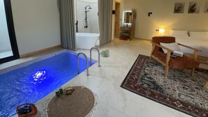 A modern hotel room with a built-in pool, a separate bathroom, a seating area, and a small dining table.