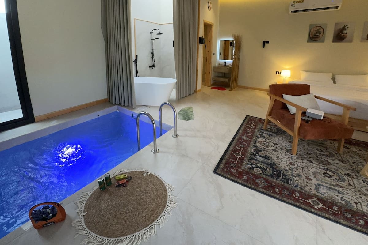 A modern hotel room with a built-in pool, a separate bathroom, a seating area, and a small dining table.
