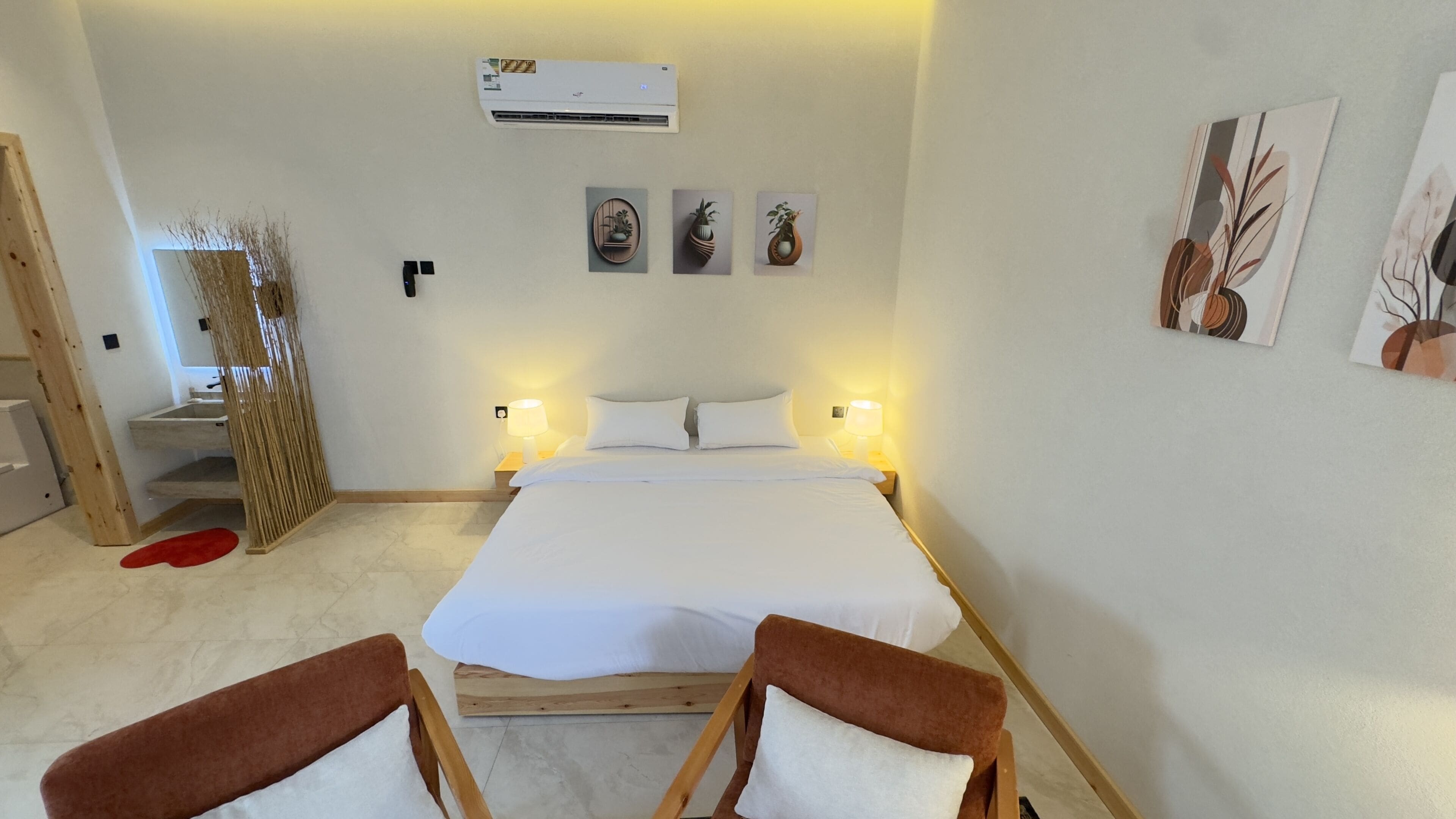 A bedroom with a bed, two bedside lamps, a wooden headboard, a red mat, and a wall-mounted air conditioner.