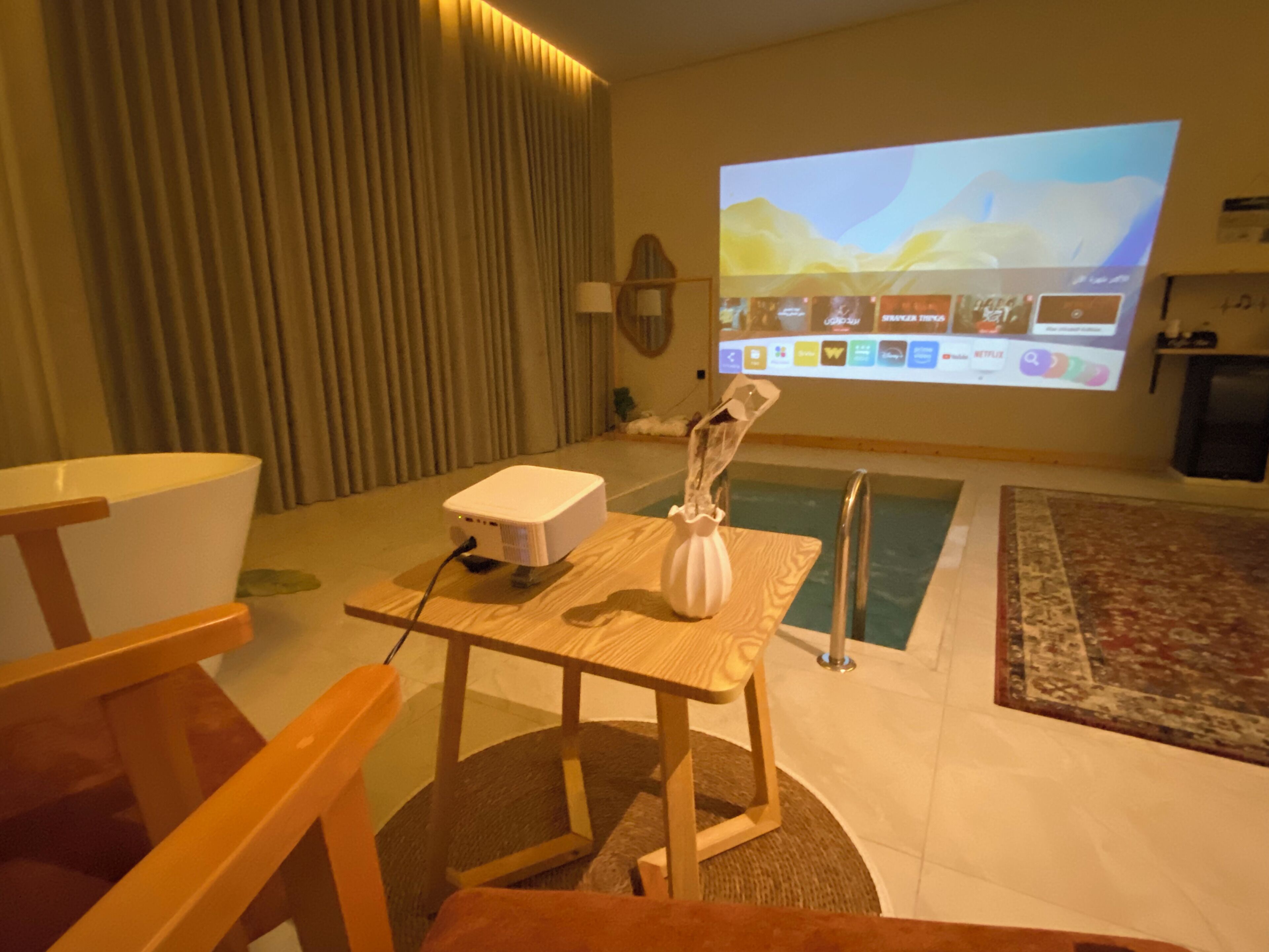 Una habitación con un proyector sobre una mesa, una pantalla grande mostrando una presentación y una piscina al fondo.