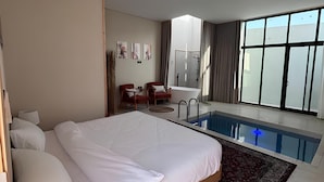 A modern hotel room with a large bed, a small seating area, a private pool, and large windows.