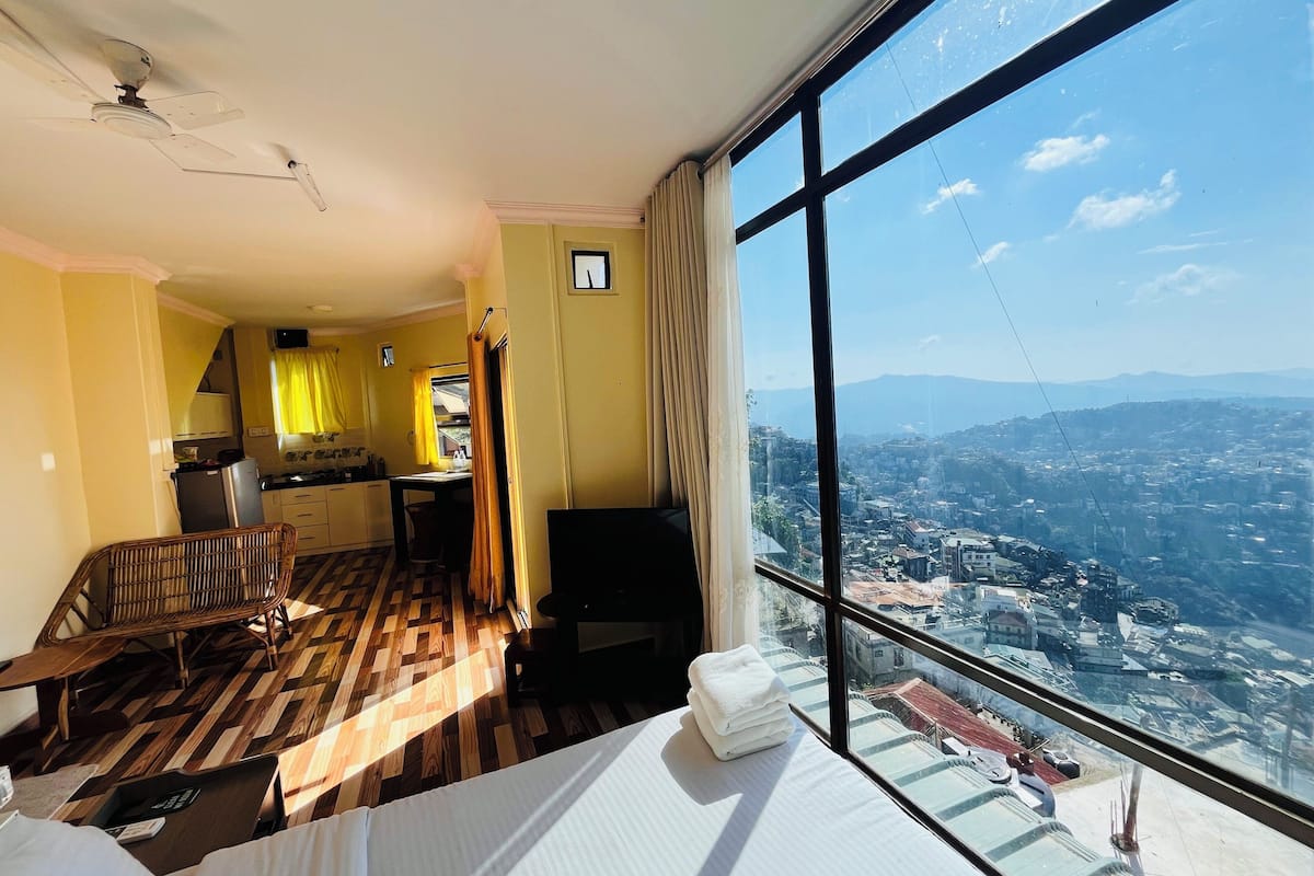 Une chambre d’hôtel avec une grande fenêtre, un lit, une chaise, une petite table et un coin cuisine.