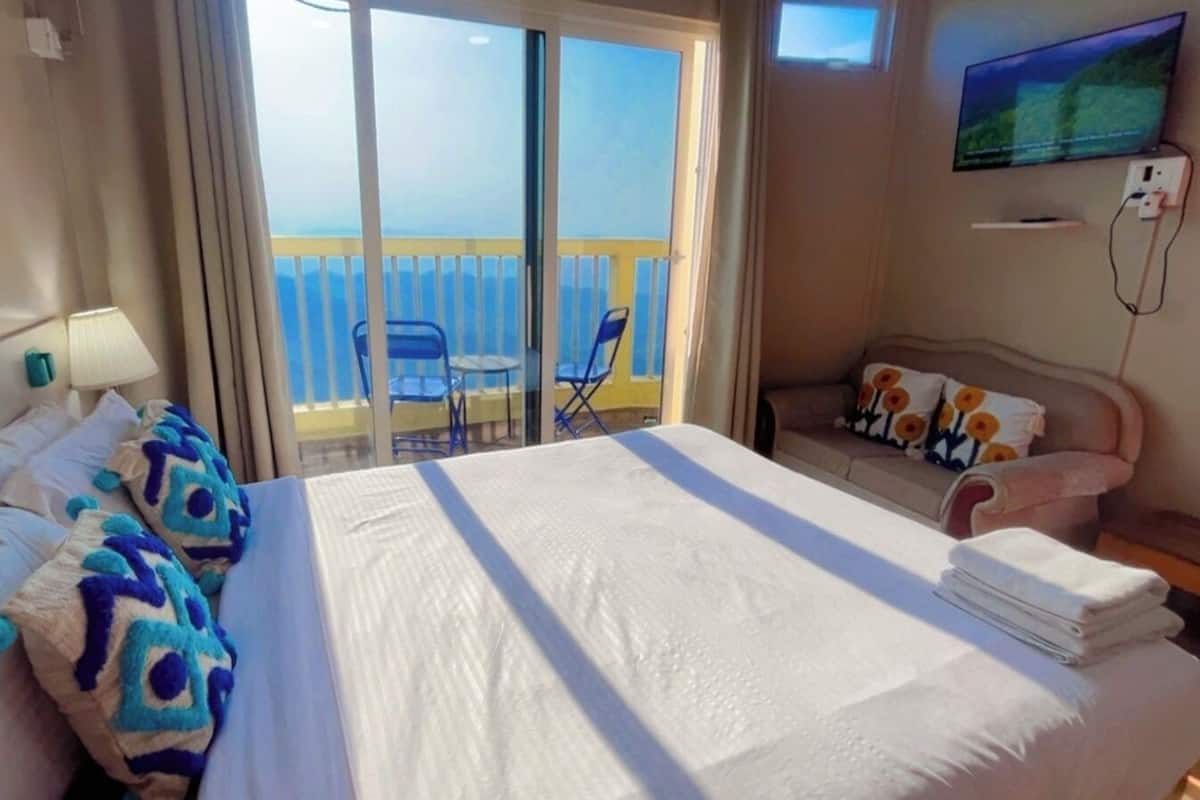 Une chambre d’hôtel avec un grand lit, une télévision, un balcon avec des chaises et une vue sur la mer.