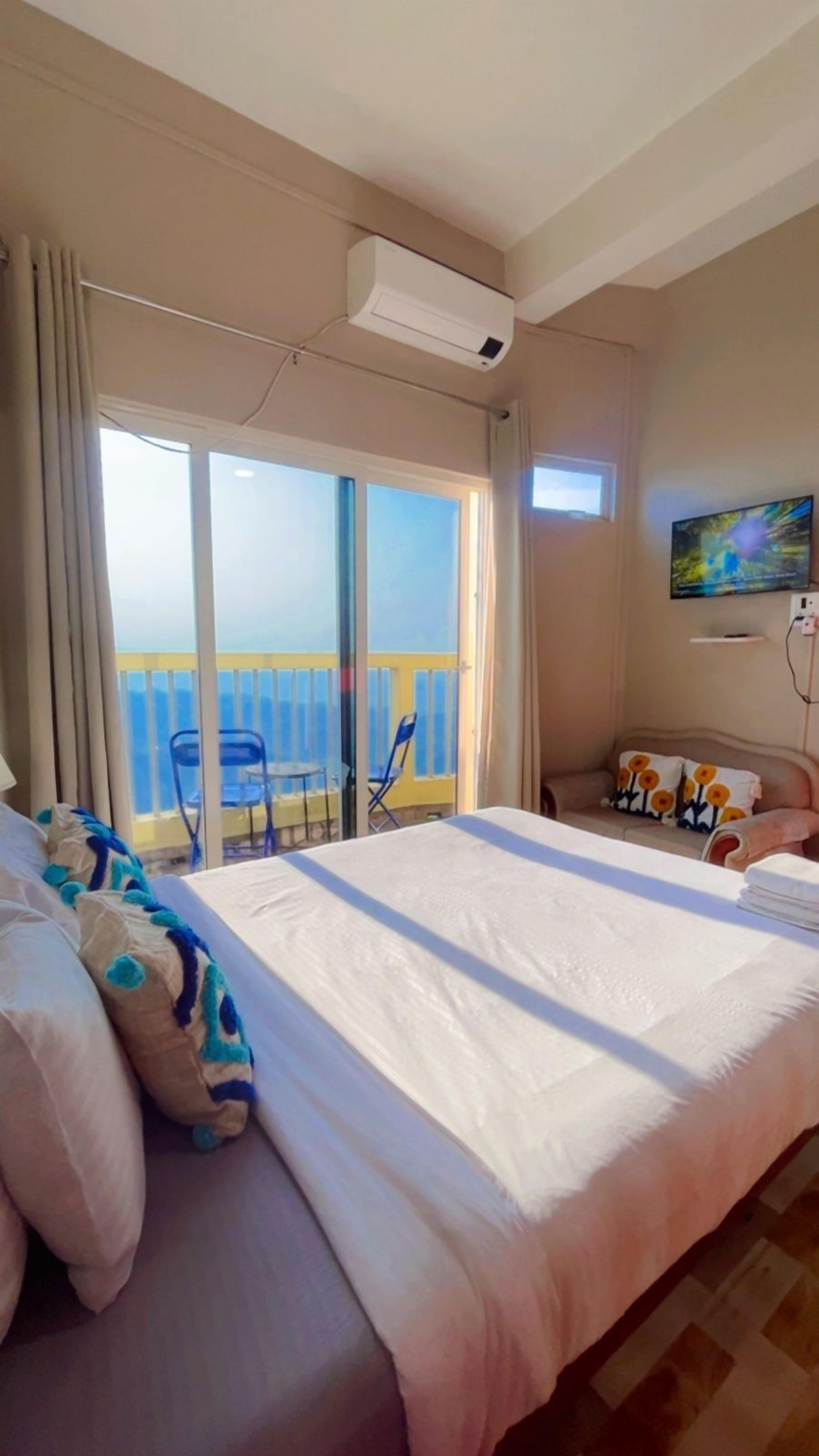 A hotel room with a large bed, a balcony with chairs, and a TV on the wall.