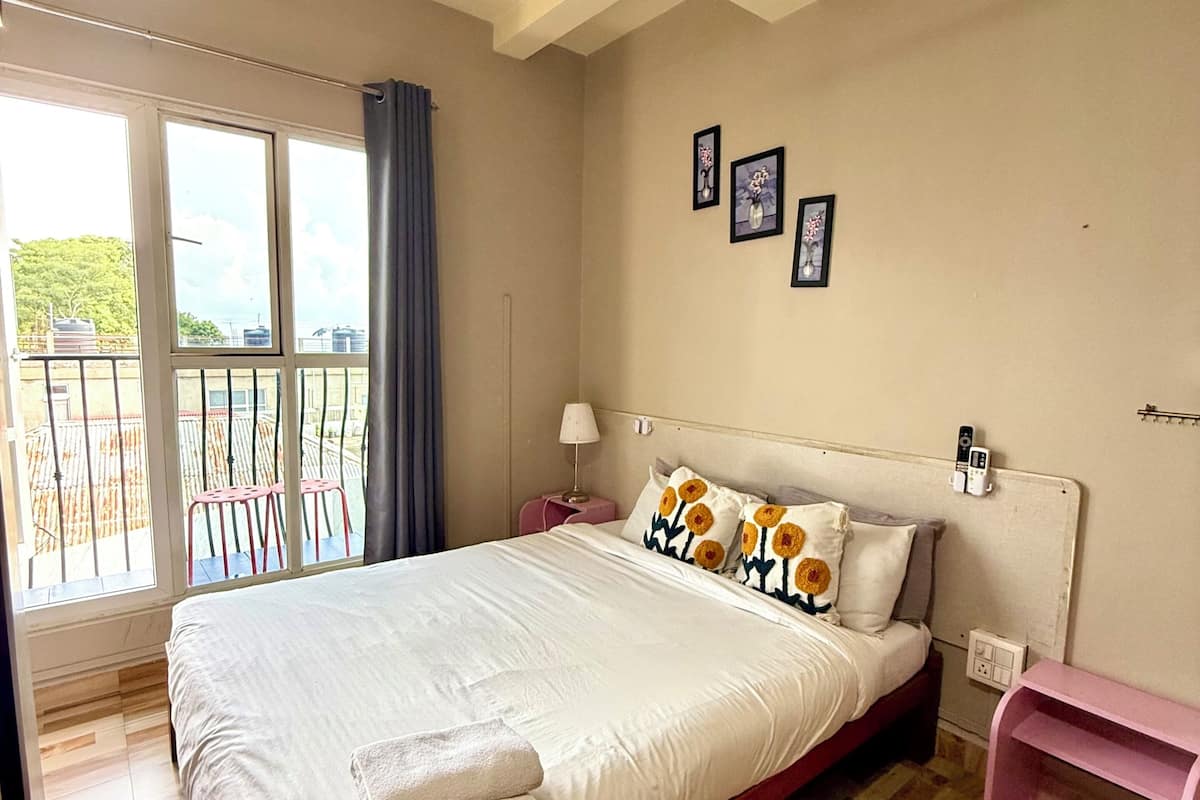 Une chambre avec un grand lit, une fenêtre donnant sur un beau paysage, une table de chevet et une étagère rose.