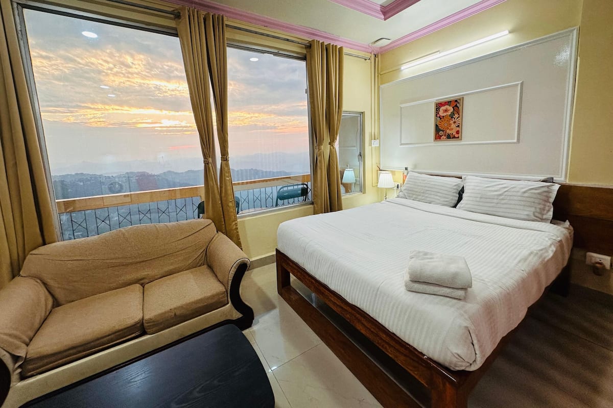 Une chambre d’hôtel avec un lit, un canapé, une fenêtre avec des rideaux, une lampe et un tableau au mur.