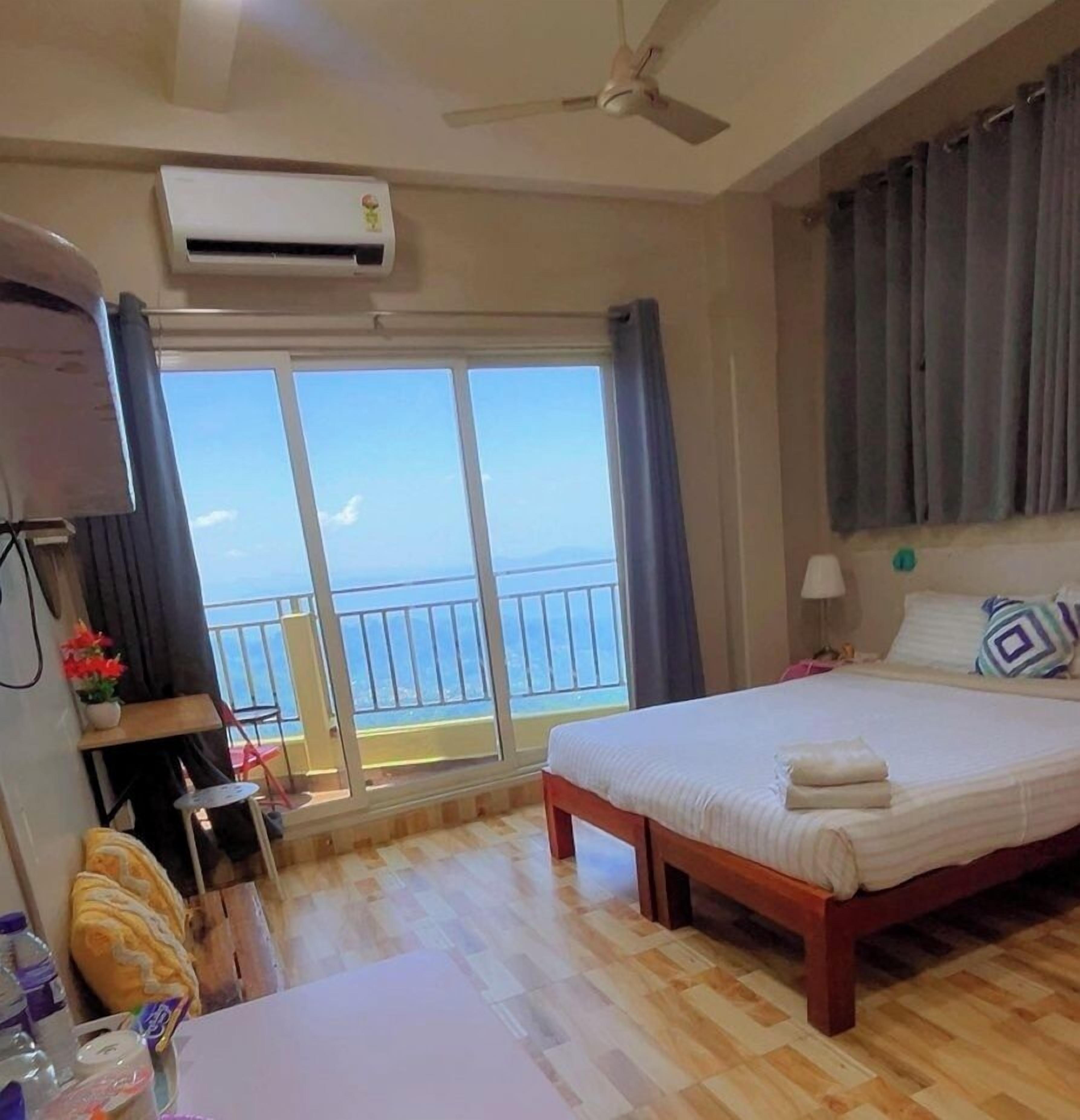 Une chambre d’hôtel avec un lit, un ventilateur de plafond, un appareil de climatisation, un balcon avec vue et une table sur laquelle se trouvent divers objets.