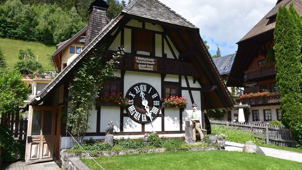 A traditional half-timbered building with a large outdoor clock, a stone statue, and flower boxes. - Semi-detached House Christa with Parking, Terrace, Private Garden and Balcony (Schonach im Schwarzwald)