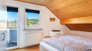 A bedroom with a wooden ceiling, a bed with checkered bedding, a white radiator, and a window with a view of trees and a wind turbine. - Semi-detached House Christa with Parking, Terrace, Private Garden and Balcony (Schonach im Schwarzwald)