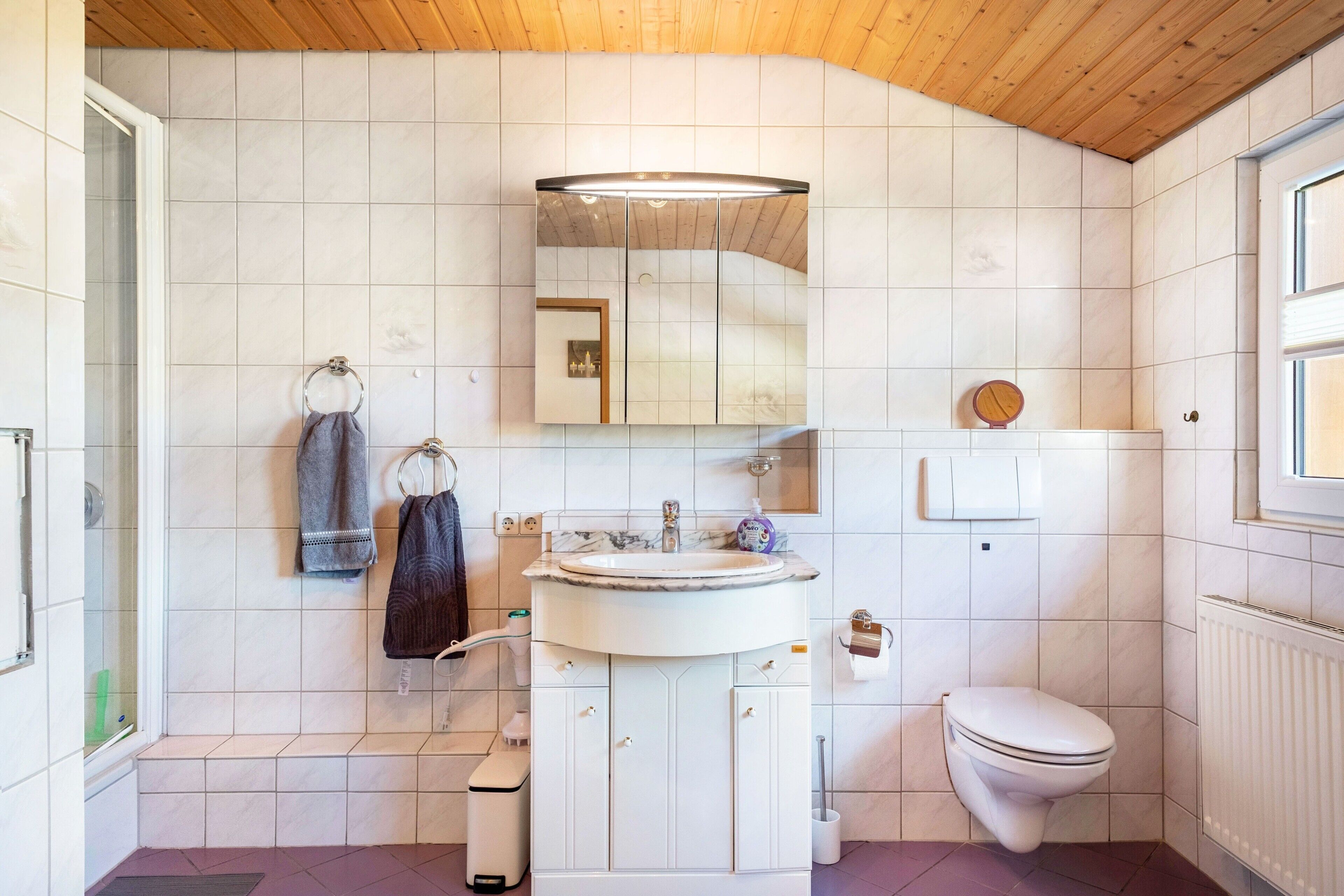 Une salle de bain avec un lavabo, un miroir et des toilettes blancs, un plafond en bois et des murs carrelés.