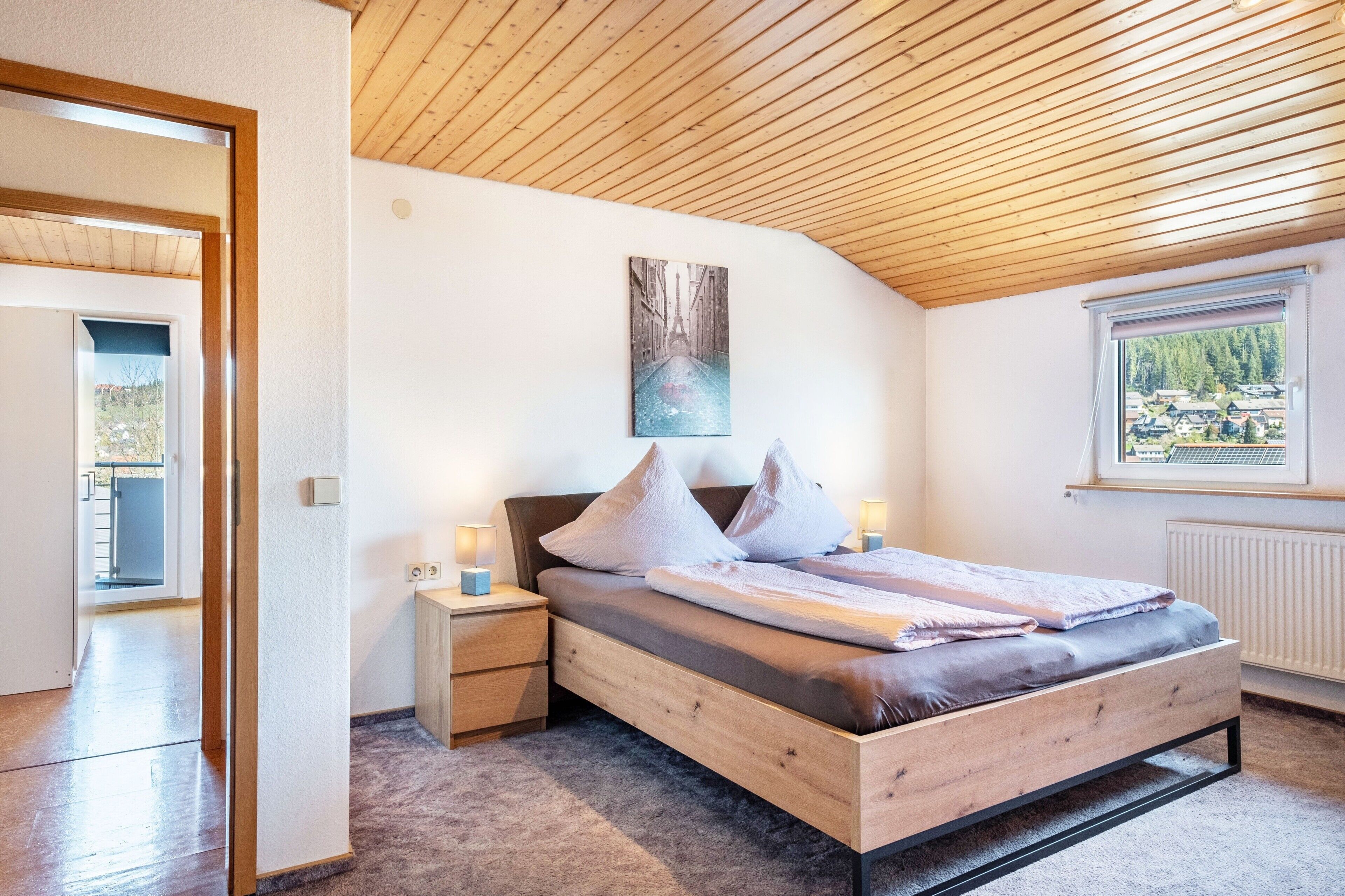 A bedroom with a wooden ceiling, a bed with pillows, a nightstand, a window with a view, and a painting on the wall.