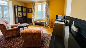 Een woonkamer met twee gestreepte banken, een kleine ronde tafel, een bankje bij het raam, en een keuken met een zwart aanrecht en spoelbak.