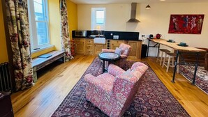 Een gezellige woonkamer met een bank, een klein rond tafeltje, een lange bank en een keuken met een aanrecht en kastjes.