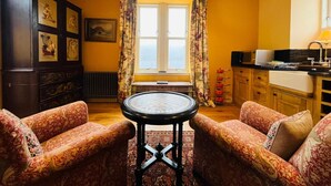 Een gezellige woonkamer met gestreepte banken, een ronde tafel, een donker kastje met kunstwerken, en een keukenhoek met raam en gordijnen.