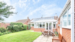 A patio area with a table and chairs, a brick house with a conservatory, and a well-maintained lawn. - Harpers - sleeps 6 guests  in 3 bedrooms (Burnham Market)