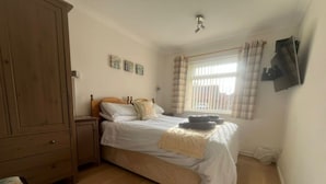 A bedroom with a wooden wardrobe, a bed with white bedding, a nightstand, a window with curtains, and a wall-mounted TV. - Harpers - sleeps 6 guests  in 3 bedrooms (Burnham Market)