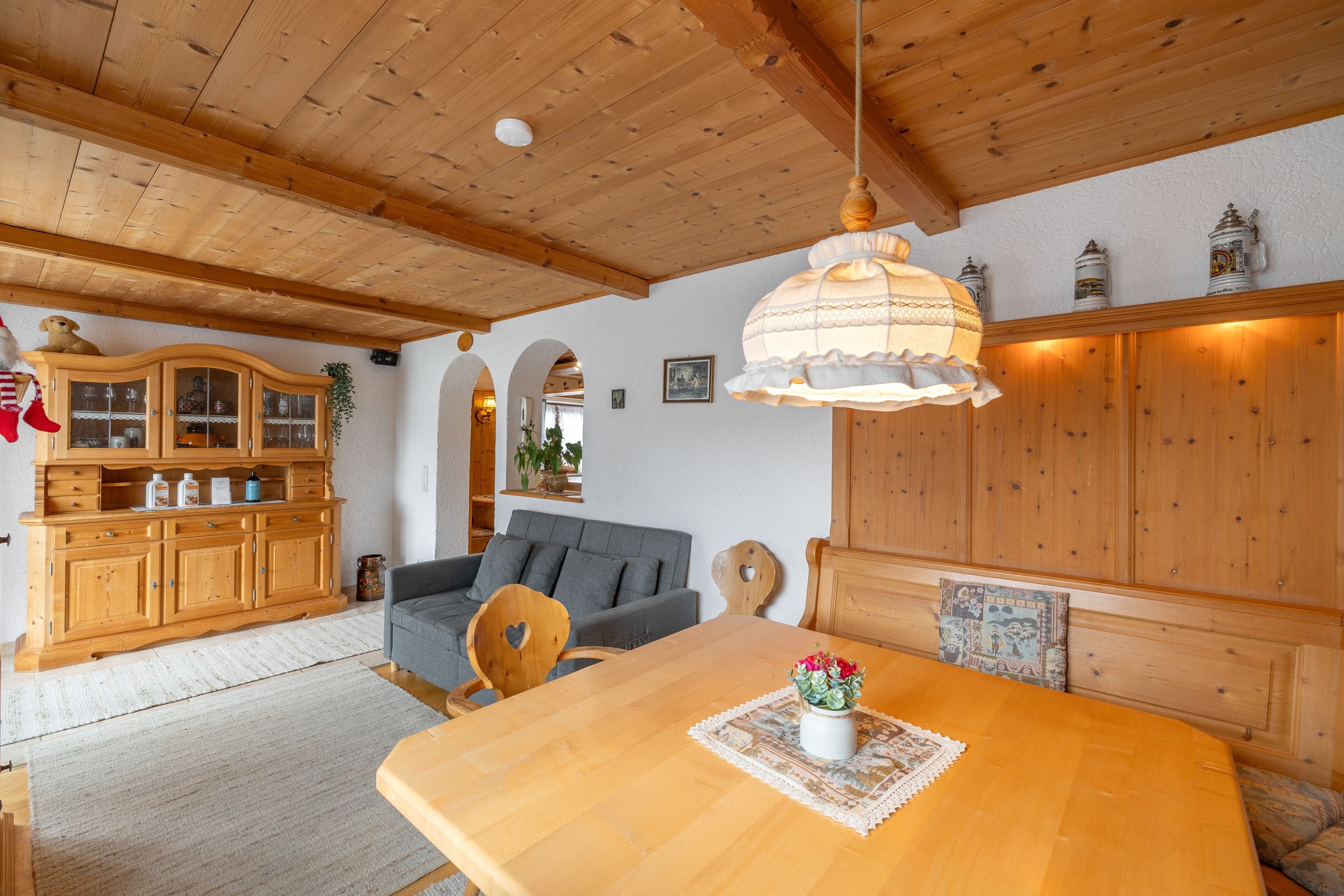 Una habitació acollidora amb bigues de fusta, una taula de dinar amb un ram de flors, un sofà i un armari amb portes de vidre.