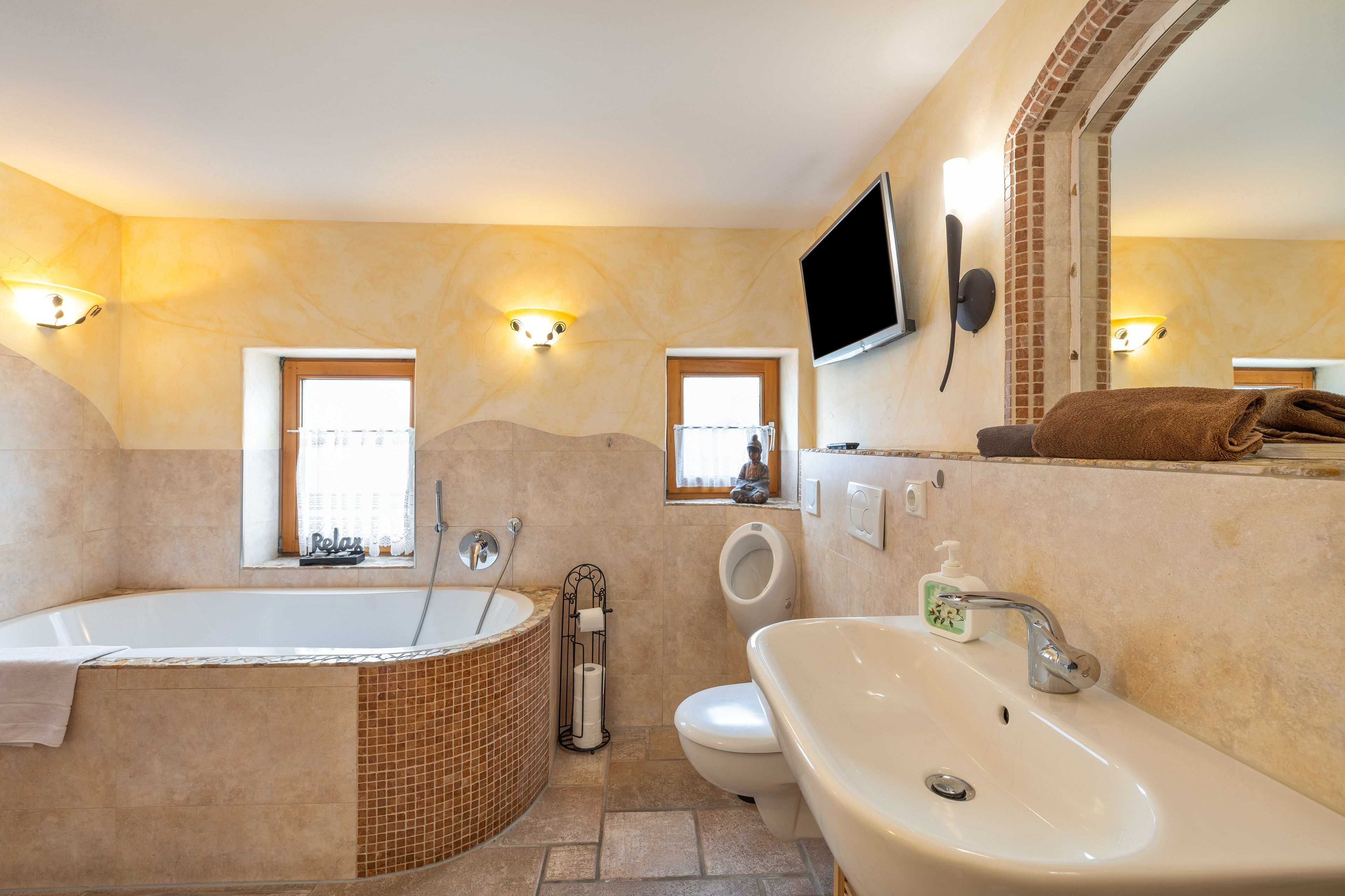 Ein Badezimmer mit einer Badewanne, einem Waschbecken, einer Toilette und einer Dusche. Zwei Fenster und ein an der Wand befestigtes Fernsehgerät.