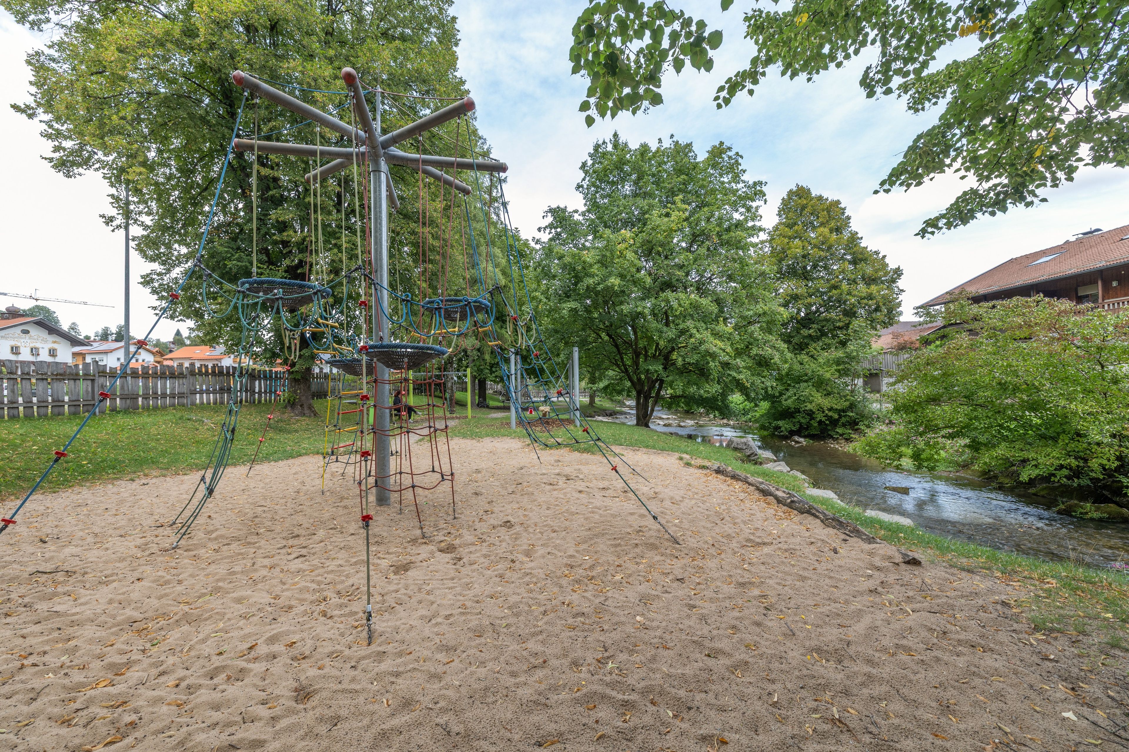 Ein Spielplatz mit Klettergerüsten und Sandbereich, umgeben von Bäumen und einem kleinen Bach.