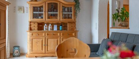 A wooden cabinet with glass doors, a dining table with a chair, and a sofa in a room with a wooden ceiling and white walls.