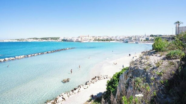 Una spiaggia costiera con acque cristalline, un litorale roccioso e una città all'orizzonte.