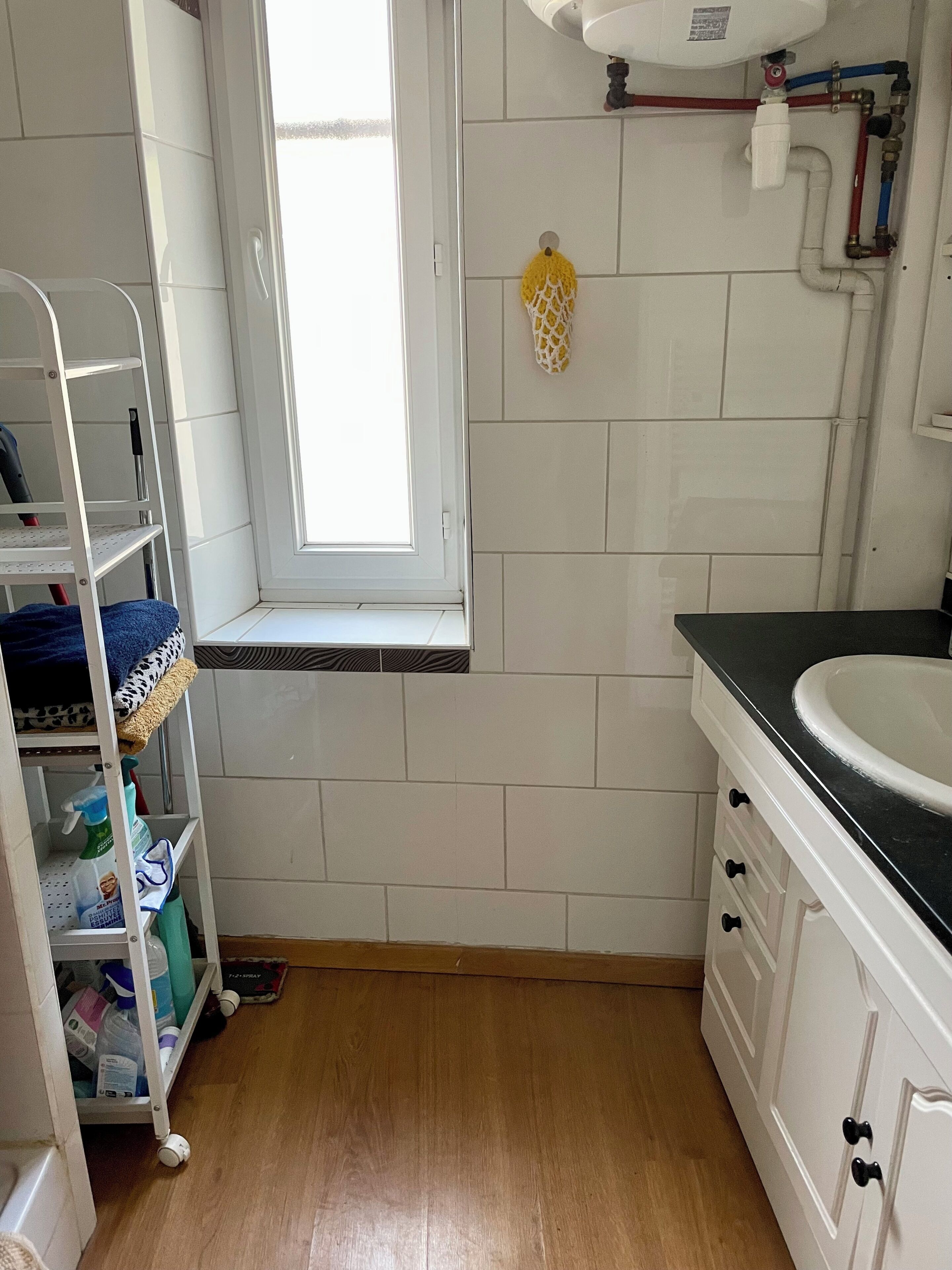 A bathroom with a window, white tiles, a sink, and a shelf with cleaning supplies.