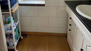 Casa de banho com janela, azulejos brancos, uma pia e um armário com produtos de limpeza.