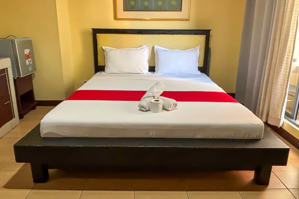 Una cama bien hecha con ropa blanca y un detalle rojo, una mesita de noche con una lámpara, y un cuadro enmarcado sobre la cama.