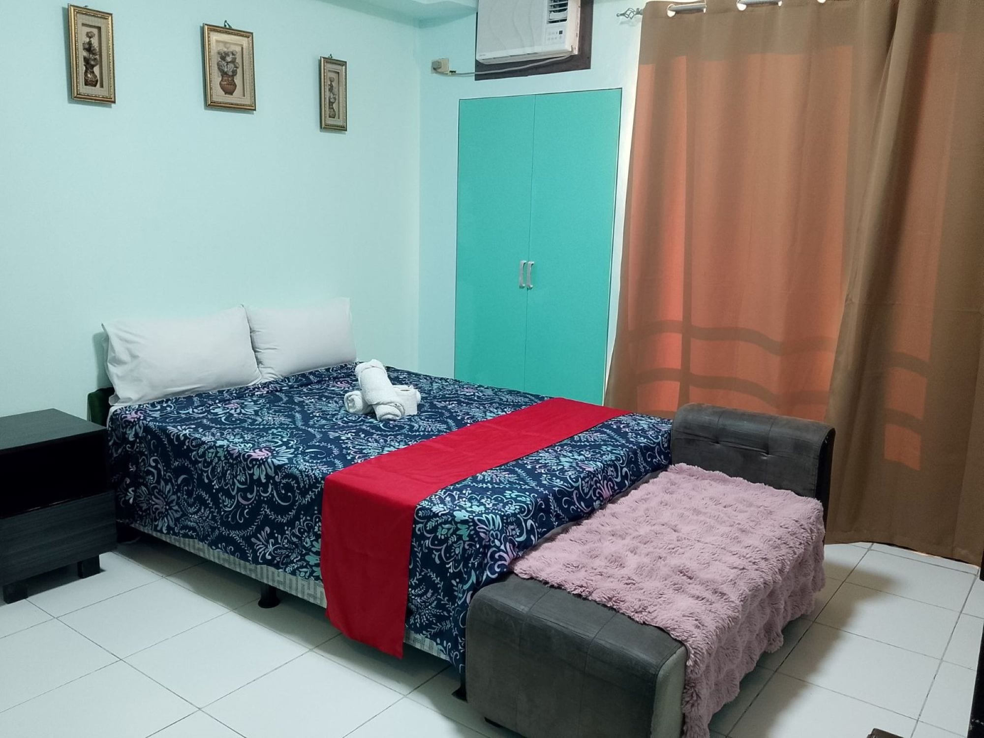 A bedroom with a bed, a nightstand, a wardrobe, a window with an air conditioner, and a curtain.