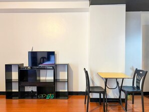Una habitación con piso de madera, un mueble negro para entretenimiento con un televisor, una mesita con dos sillas y una pared blanca.