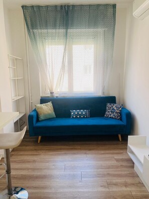 Ein Zimmer mit einem blauen Sofa, einem weißen Regal und einem Fenster mit durchsichtigen Vorhängen.