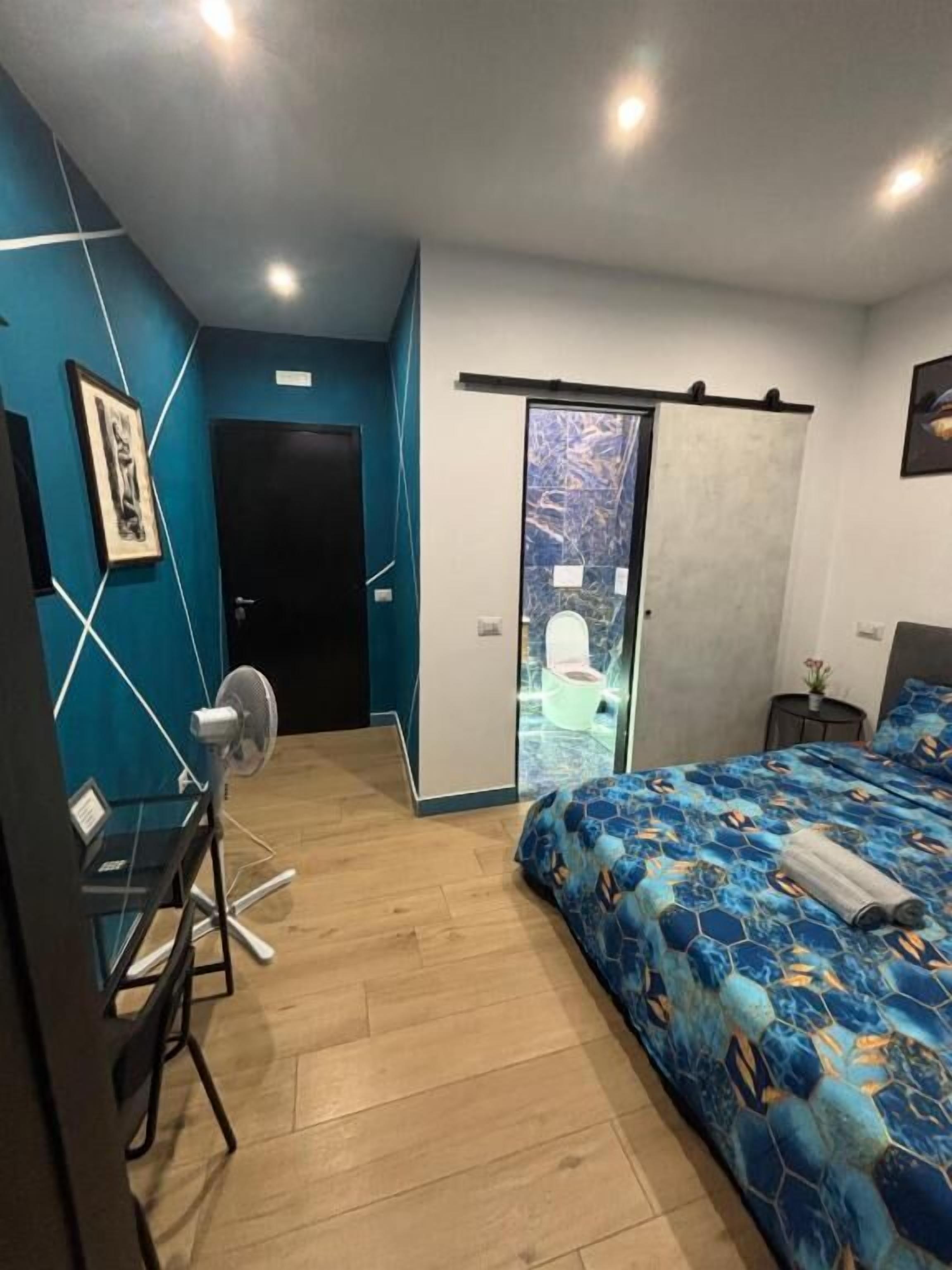 A bedroom with a bed, a desk, a chair, a fan, a door, and a view of the outside.