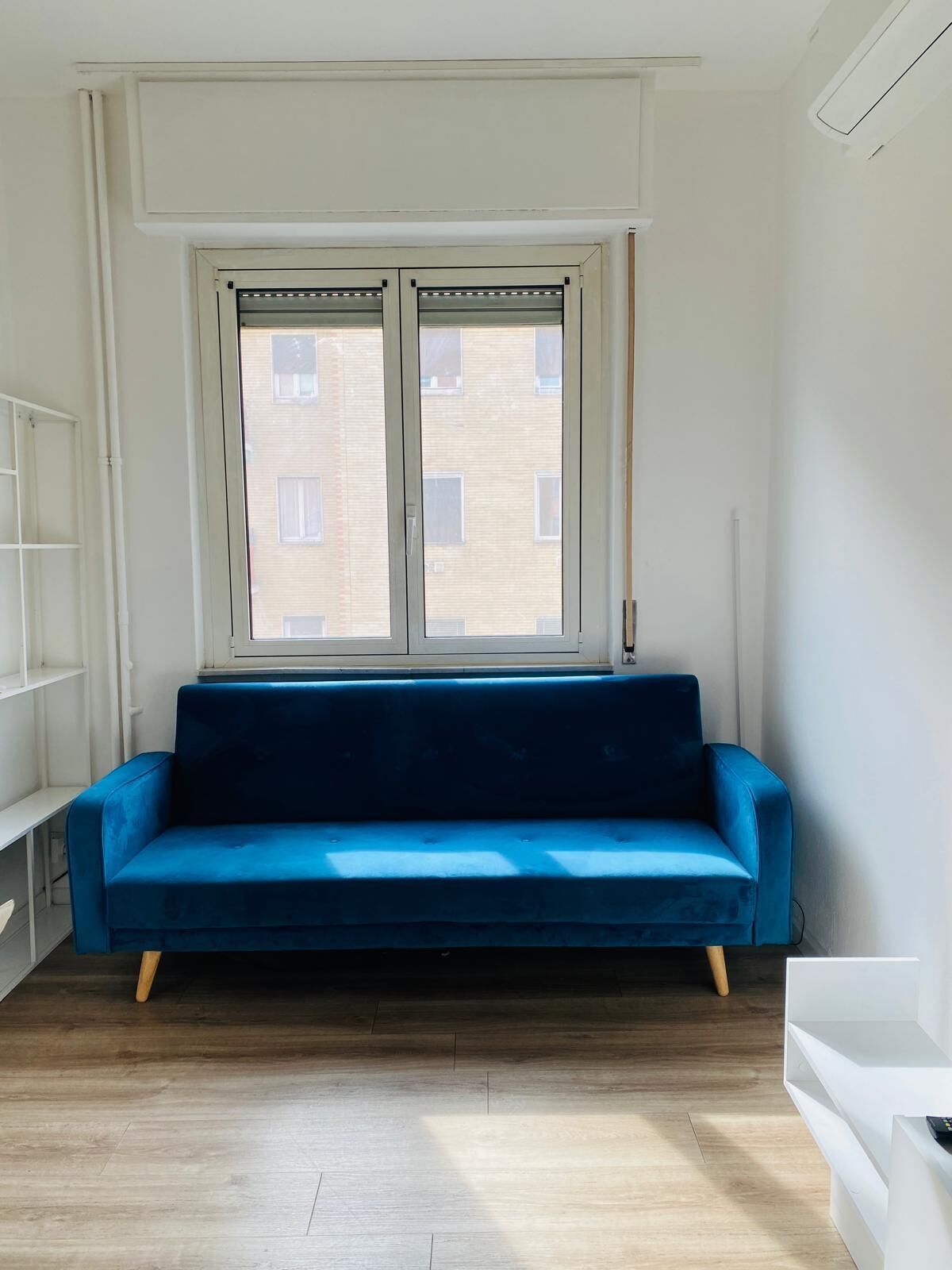 A room with a blue sofa, a window with blinds, a white radiator, and a white shelf unit.