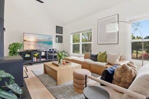 Una sala moderna con sofá, mesa de centro y televisor, una ventana grande con vistas, y un mueble de entretenimiento negro.